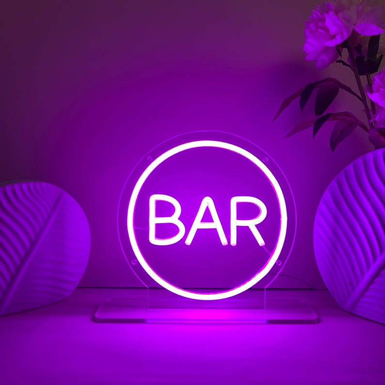 May include: A neon sign with the word "BAR" in white letters inside a white circular frame. The sign is illuminated with a bright purple light and sits on a clear stand. Decorative vases and flowers are in the background.