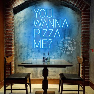 May include: A bright blue neon sign mounted on a wall displaying the text YOU WANNA PIZZA ME? in a modern sans-serif font. The sign is positioned above a wooden table with two chairs in a room featuring exposed brick archway architectural details.