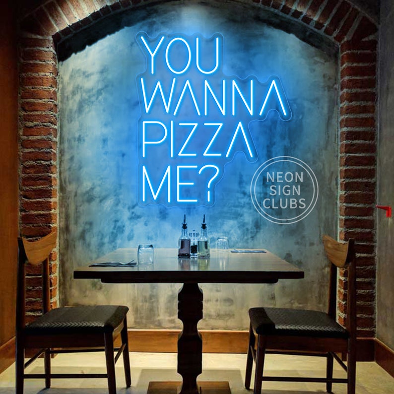 May include: A bright blue neon sign mounted on a wall displaying the text YOU WANNA PIZZA ME? in a modern sans-serif font. The sign is positioned above a wooden table with two chairs in a room featuring exposed brick archway architectural details.