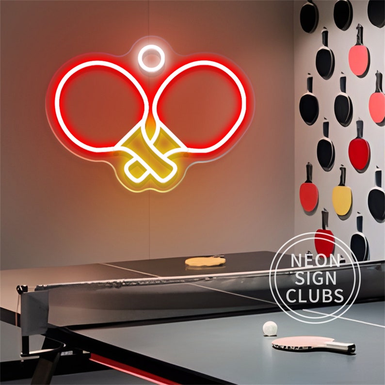 May include: A neon sign in the shape of two ping pong paddles with a ball above them, glowing red and yellow. A ping pong table and paddle are in the foreground. The sign is above a ping pong table.