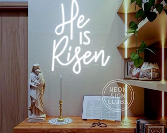 Könnte beinhalten: Ein weißes Neon-Schild mit den Worten "He is Risen" an einer beigefarbenen Wand. Eine Statue von Maria und Jesus steht auf einem Holztisch mit einer Kerze und einem Buch.