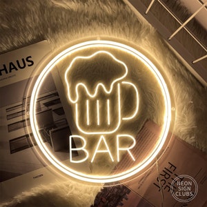 May include: A round, illuminated neon sign with a beer mug graphic and the word "BAR" in white. The sign is surrounded by a circular frame and emits a warm, yellow glow. The sign is from Neon Sign Clubs.