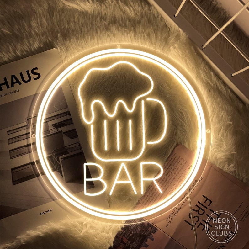 May include: A round, illuminated neon sign with a beer mug graphic and the word "BAR" in white. The sign is surrounded by a circular frame and emits a warm, yellow glow. The sign is from Neon Sign Clubs.