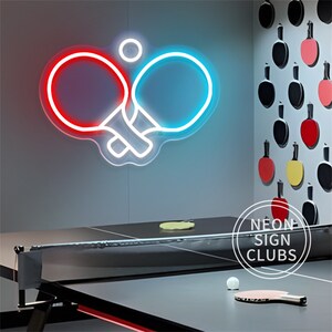 May include: A neon sign featuring two crossed ping pong paddles, one red and one blue, with a white outline. A white ping pong ball is above the paddles. A ping pong table and paddles are in the foreground. The text "NEON SIGN CLUBS" is in a white circle.