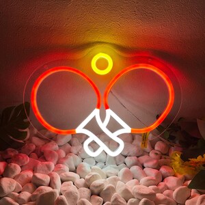 May include: A neon sign in the shape of two ping pong paddles with a yellow circle above. The paddles are outlined in red with white handles. The sign is placed on a bed of white stones.