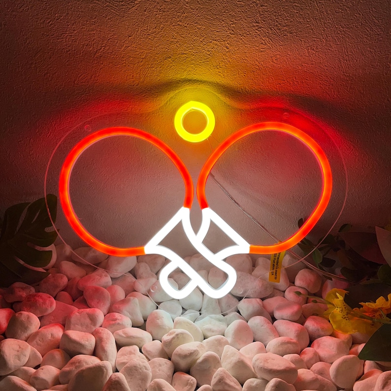 May include: A neon sign in the shape of two ping pong paddles with a yellow circle above. The paddles are outlined in red with white handles. The sign is placed on a bed of white stones.