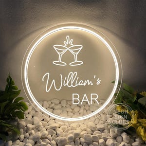 Peut inclure: Panneau lumineux rond avec l'inscription "William's BAR" en cursive et deux verres à cocktail. Le signe émet une lueur blanche et est placé sur un fond de galets blancs et de verdure.