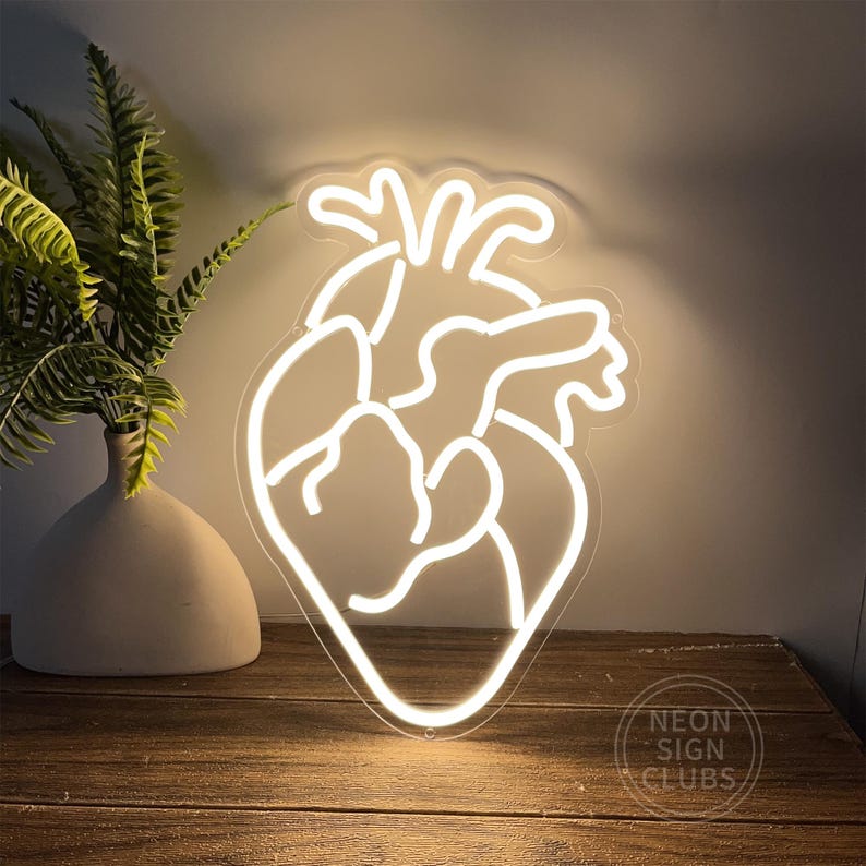 Peut inclure: Une enseigne au n&eacute;on blanche en forme de c&oelig;ur humain, avec le texte "Neon Sign Clubs" en bas &agrave; droite. L'enseigne est pos&eacute;e sur une surface en bois, avec un vase et une plante &agrave; gauche.