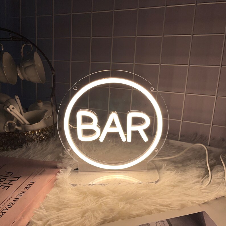 May include: A white neon "BAR" sign in a clear circular frame, mounted on a white base. The sign is illuminated, casting a soft glow. The background includes a fluffy white surface and a tiled wall.