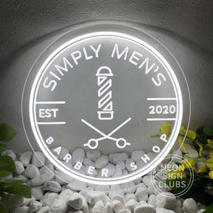 Może przedstawiać: Okrągły biały neonowy szyld dla zakładu fryzjerskiego z napisem "SIMPLY MEN'S BARBER SHOP". Projekt zawiera znak fryzjerski, nożyczki i rok "2020". Znak otoczony jest białymi kamieniami i zielenią.