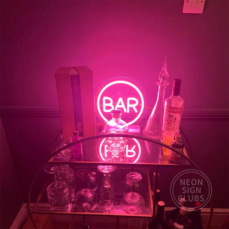 May include: A pink neon sign that says "BAR" in a circle, displayed on a bar cart with bottles, glasses, and a skull-shaped bottle. The cart has two glass shelves and a gold metal frame. The background is a dark wall.