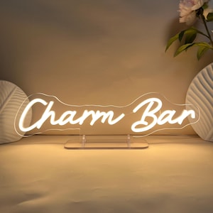 May include: A neon sign displaying the words "Charm Bar" in a cursive script. The sign emits a warm white light and is mounted on a clear acrylic base. The background is a neutral beige colour.