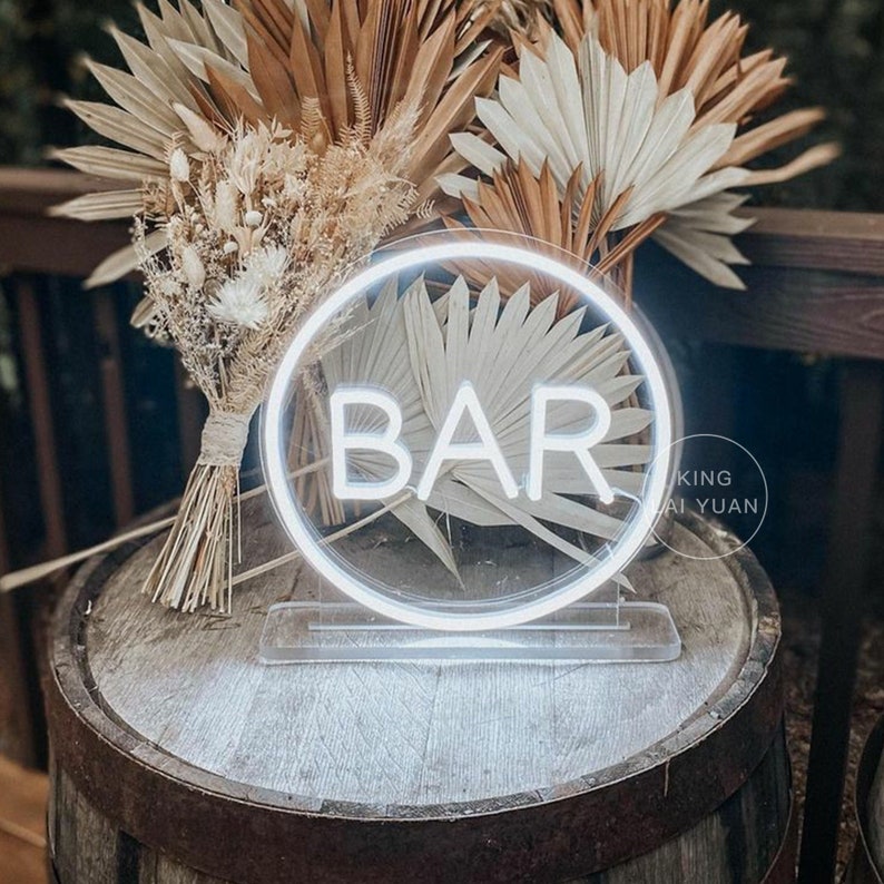 May include: A white neon "BAR" sign in a circular frame, resting on a clear stand. The sign is placed on a wooden barrel, with dried floral arrangements in the background. The sign is illuminated with a bright white light.