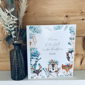 Op de afbeelding: Een wit boek met een groene en blauwe rand. Het boek heeft een titel die "Meine Kita Zeit in der Fridoline" luidt en de naam "Sarah". Het boek is versierd met cartoondieren die hoofdtooien dragen.