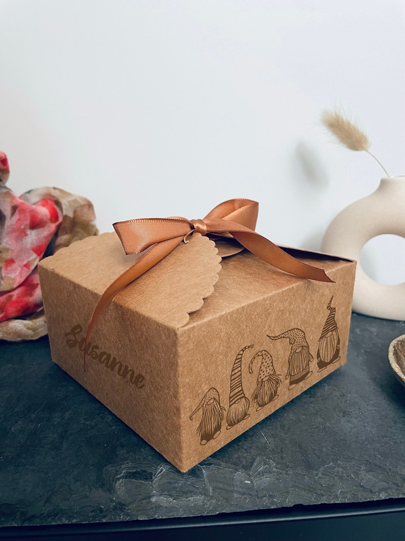 Puede incluir: Una caja de regalo de cart&oacute;n marr&oacute;n atada con una cinta de color cobre. La caja presenta un borde festoneado y un dise&ntilde;o grabado con l&aacute;ser de gnomos. El nombre "Susanne" est&aacute; grabado en el lateral.