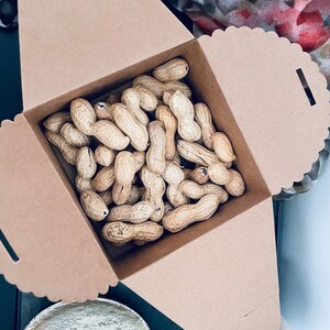 Puede incluir: Una caja de cart&oacute;n marr&oacute;n llena de cacahuetes sin c&aacute;scara. La caja tiene un borde festoneado y est&aacute; parcialmente abierta, revelando los cacahuetes en el interior. Los cacahuetes son de color beige y tienen una textura rugosa.