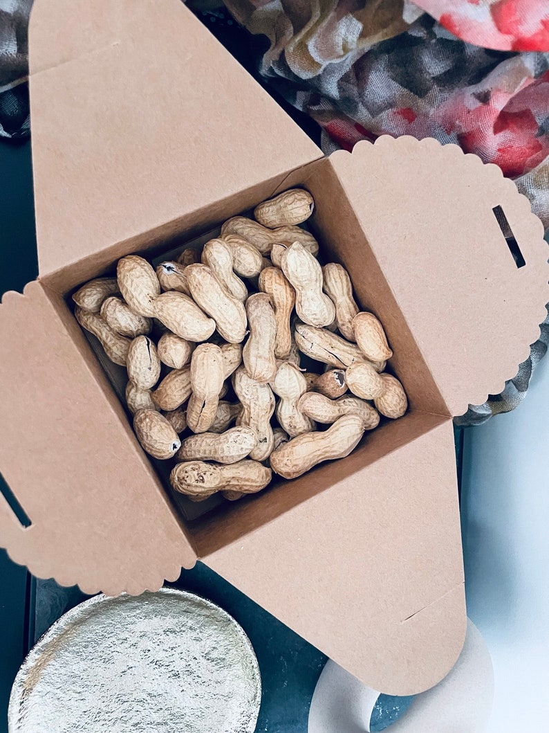 Puede incluir: Una caja de cart&oacute;n marr&oacute;n llena de cacahuetes sin c&aacute;scara. La caja tiene un borde festoneado y est&aacute; parcialmente abierta, revelando los cacahuetes en el interior. Los cacahuetes son de color beige y tienen una textura rugosa.