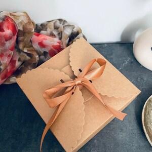 Puede incluir: Una peque&ntilde;a caja de regalo cuadrada con bordes festoneados de cart&oacute;n marr&oacute;n, atada con una cinta de raso color melocot&oacute;n. Una bufanda floral con tonos rojos y marrones es parcialmente visible en el fondo.
