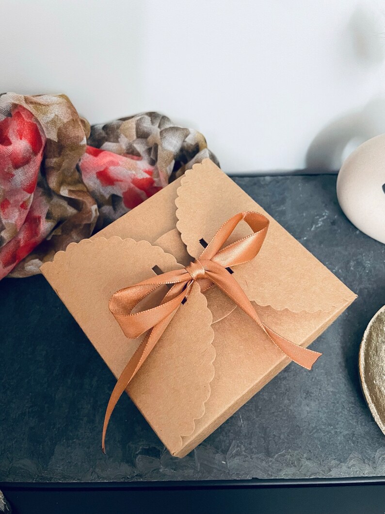 Puede incluir: Una peque&ntilde;a caja de regalo cuadrada con bordes festoneados de cart&oacute;n marr&oacute;n, atada con una cinta de raso color melocot&oacute;n. Una bufanda floral con tonos rojos y marrones es parcialmente visible en el fondo.