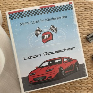 Könnte beinhalten: Ein blauer und roter Ordner mit einem karierten Muster und dem Text "Meine Zeit im Kindergarten" und "Leon Rauscher". Der Ordner zeigt ein rotes Rennwagen und einen roten Rennhelm mit einer karierten Flagge.
