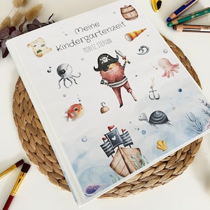 Op de afbeelding: Een wit fotoalbum met een blauwe en witte aquarel-illustratie van een piratenschip, een octopus, een schildpad en een schatkist. De tekst "Meine Kindergartenzeit" en "MORITZ STEPHAN" staat op de omslag gedrukt.