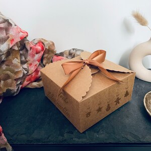 Puede incluir: Una peque&ntilde;a caja de regalo de cart&oacute;n marr&oacute;n atada con una cinta beige. La caja tiene un borde festoneado y est&aacute; decorada con ilustraciones navide&ntilde;as y las palabras "Frohe Weihnachten". Una bufanda decorativa y un jarr&oacute;n est&aacute;n en el fondo.