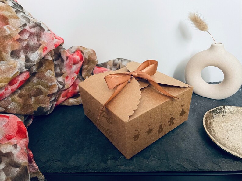 Puede incluir: Una peque&ntilde;a caja de regalo de cart&oacute;n marr&oacute;n atada con una cinta beige. La caja tiene un borde festoneado y est&aacute; decorada con ilustraciones navide&ntilde;as y las palabras "Frohe Weihnachten". Una bufanda decorativa y un jarr&oacute;n est&aacute;n en el fondo.