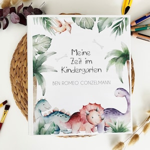 Op de afbeelding: Een wit boek met een groene, blauwe en paarse dinosaurus illustratie. Het boek heeft de tekst "Meine Zeit im Kindergarten" en "BEN ROMEO CONZELMANN" op de voorkant gedrukt.