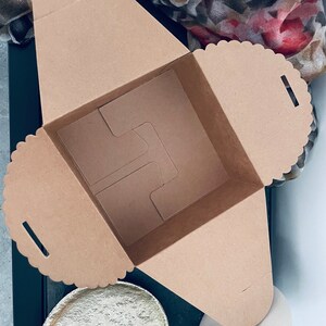 Puede incluir: Caja de cart&oacute;n marr&oacute;n cuadrada y abierta, con borde festoneado. Contiene un inserto interno para sujetar objetos. Sobre una superficie blanca, con un plato dorado y un jarr&oacute;n blanco.
