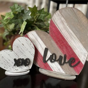Könnte beinhalten: Zwei herzförmige Schilder aus Holz mit einer abgenutzten weißen und roten Lackierung. Das größere Schild sagt "love" in schwarzer Schrift. Das kleinere Schild sagt "xoxo" in schwarzer Schrift.
