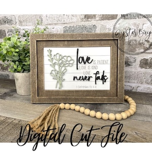 May include: A framed wood sign with a white background and black text that reads "Love is patient, Love is kind, Love never fails, 1 Corinthians 13:4-8". The sign also features a floral design with a green stem and white flowers.