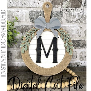 May include: A wooden round sign with a gray bow, green leaves, and the letter "M" in black on a white background. The sign has a wooden handle on the top. The text "Digital Cut File" is visible at the bottom of the image.