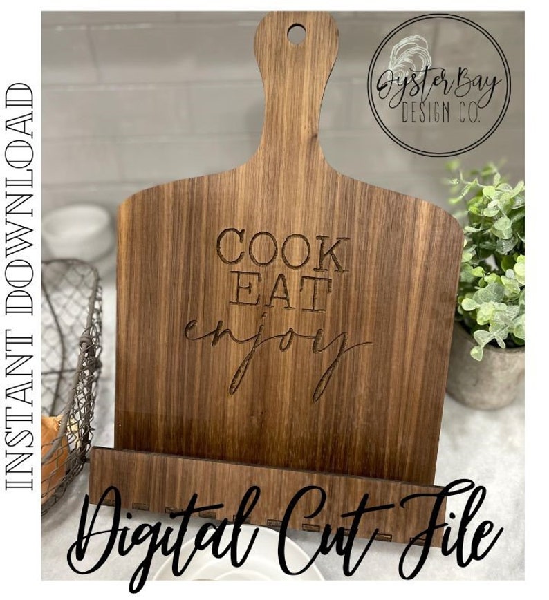 K&ouml;nnte beinhalten: Holzschneidebrett mit Griff und eingebautem St&auml;nder. Auf dem Brett stehen die Worte "COOK EAT enjoy". Die Worte "Instant Download" und "Digital Cut File" sind ebenfalls sichtbar. Das Brett ist braun und hat eine nat&uuml;rliche Holzmaserung.