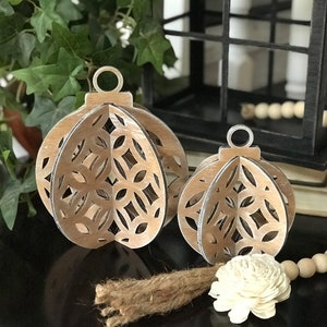 May include: Two wooden ornaments with a whitewash finish and intricate cut-out designs. The ornaments are shaped like spheres and have a rustic, farmhouse style.
