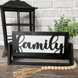 May include: Black wooden sign with the word "family" in white lettering on a white and black chevron background. The sign is on a black stand with a wooden bead garland and a jute tassel.