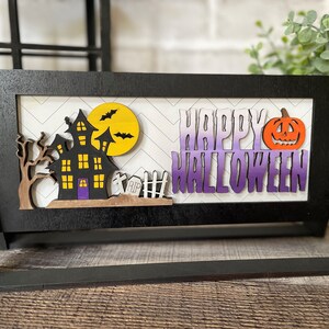 May include: A black wooden Halloween-themed sign with a haunted house, bats, and a pumpkin. The words "HAPPY HALLOWEEN" are displayed in purple and white. The sign is on a wooden surface.