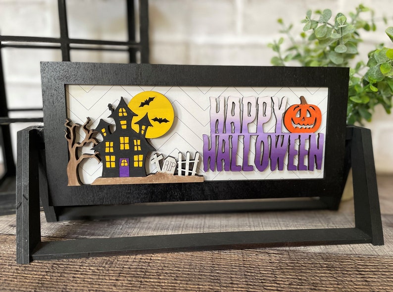 May include: A black wooden Halloween-themed sign with a haunted house, bats, and a pumpkin. The words "HAPPY HALLOWEEN" are displayed in purple and white. The sign is on a wooden surface.