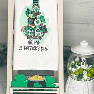Puede incluir: Toalla de cocina blanca con una ilustración verde, negra y blanca de un puesto de varios niveles con un duende, tréboles y una olla de oro. La toalla dice "Happy St. Patrick's Day". Debajo de la toalla hay un estante de madera con una olla de oro y tréboles.