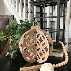 May include: A wooden, laser-cut ornament shaped like a Christmas tree bauble with a geometric pattern. The ornament is painted gold and has a jute tassel and a white sola flower attached to it. The ornament is sitting on a black table with a wooden bead garland.