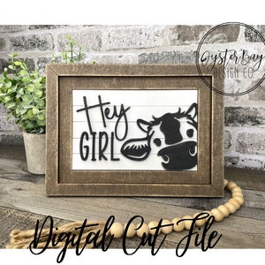 May include: A wooden framed sign with the text "Hey Girl" and a black and white cow peeking out from the right side of the frame. The sign is on a wooden surface with a strand of wooden beads.
