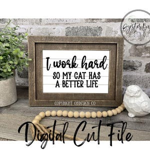 May include: A framed sign with the text "I work hard so my cat has a better life." The sign is in a brown wooden frame and sits on a wooden surface. A white bird figurine and wooden beads are also visible.
