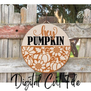 May include: A round wooden sign with the words "hey PUMPKIN" in black and orange. The bottom half features a laser-cut design of pumpkins and sunflowers. The sign is hanging on a wooden fence.