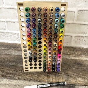 May include: A wooden stand with 72 holes for storing markers. The stand is made of light wood and has a natural finish. The markers are arranged in rows and columns, and each marker has a number on it. The stand is labeled "TOOLI-ART" and a marker is labeled "Acrylic Paint Pen".