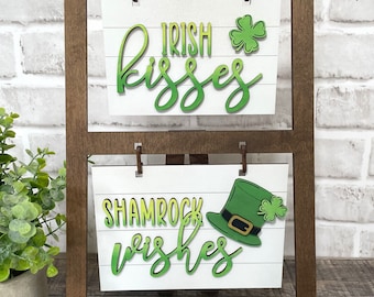 May include: A wooden easel with two white signs. The top sign says "Irish Kisses" with a green shamrock. The bottom sign says "Shamrock Wishes" with a green leprechaun hat and shamrock.