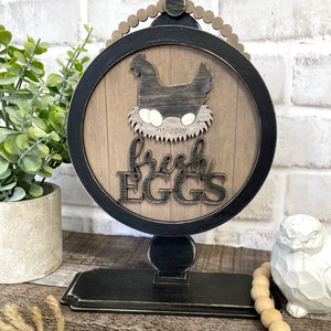 May include: Black and brown wooden sign with a black chicken silhouette sitting on a nest of eggs. The sign reads "fresh EGGS" and is on a black stand with a round base.