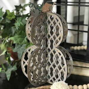 May include: A decorative wooden pumpkin with a lattice design, painted in a light brown and white wash. The pumpkin is standing on a black surface with a brown rope and a white flower.