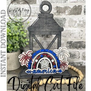 May include: A wooden cutout of a rainbow with red, white, and blue stripes, with the word "america" in the center. The rainbow is decorated with fireworks on either side. The cutout is sitting on a wooden lantern.