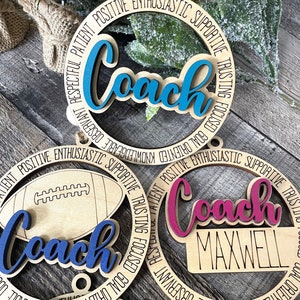 Peut inclure: Trois ornements en bois avec le mot "Coach" en diff&eacute;rentes couleurs. Les ornements sont d&eacute;cor&eacute;s d'un ballon de football, d'un ballon de basket-ball et d'un ballon de football. Le texte autour des ornements est "Respectueux, Patient, Positif, Enthousiaste, Soutenu, Confiant, Concentr&eacute;, Orient&eacute; vers les objectifs, Connaissant, Observable, Respectueux".