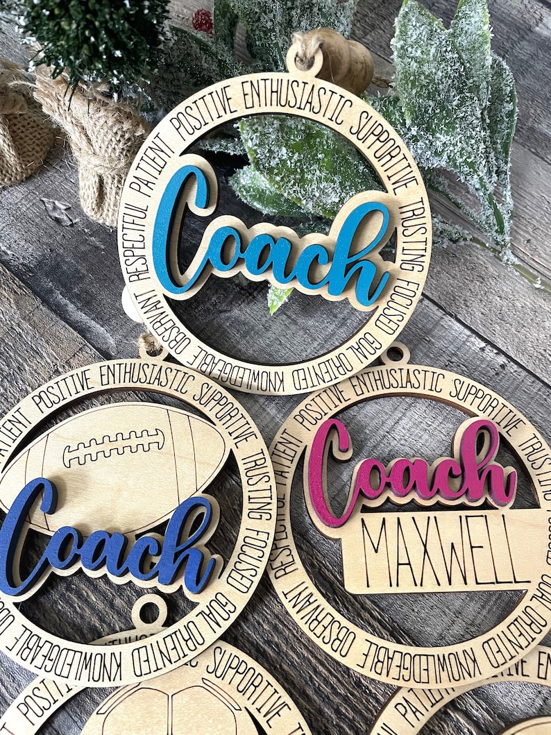 Peut inclure: Trois ornements en bois avec le mot "Coach" en diff&eacute;rentes couleurs. Les ornements sont d&eacute;cor&eacute;s d'un ballon de football, d'un ballon de basket-ball et d'un ballon de football. Le texte autour des ornements est "Respectueux, Patient, Positif, Enthousiaste, Soutenu, Confiant, Concentr&eacute;, Orient&eacute; vers les objectifs, Connaissant, Observable, Respectueux".