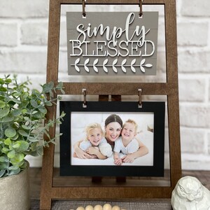 May include: A wooden easel with a gray sign that says "simply blessed" and a black frame with a photo of a mother and her two children. The easel is on a wooden surface with a string of wooden beads and a white owl figurine.